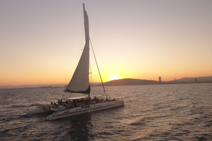 Sailboat on the water at sunset with city skyline and hills in background.