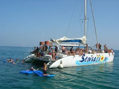 People on a catamaran with some swimming nearby in the ocean on a clear day.